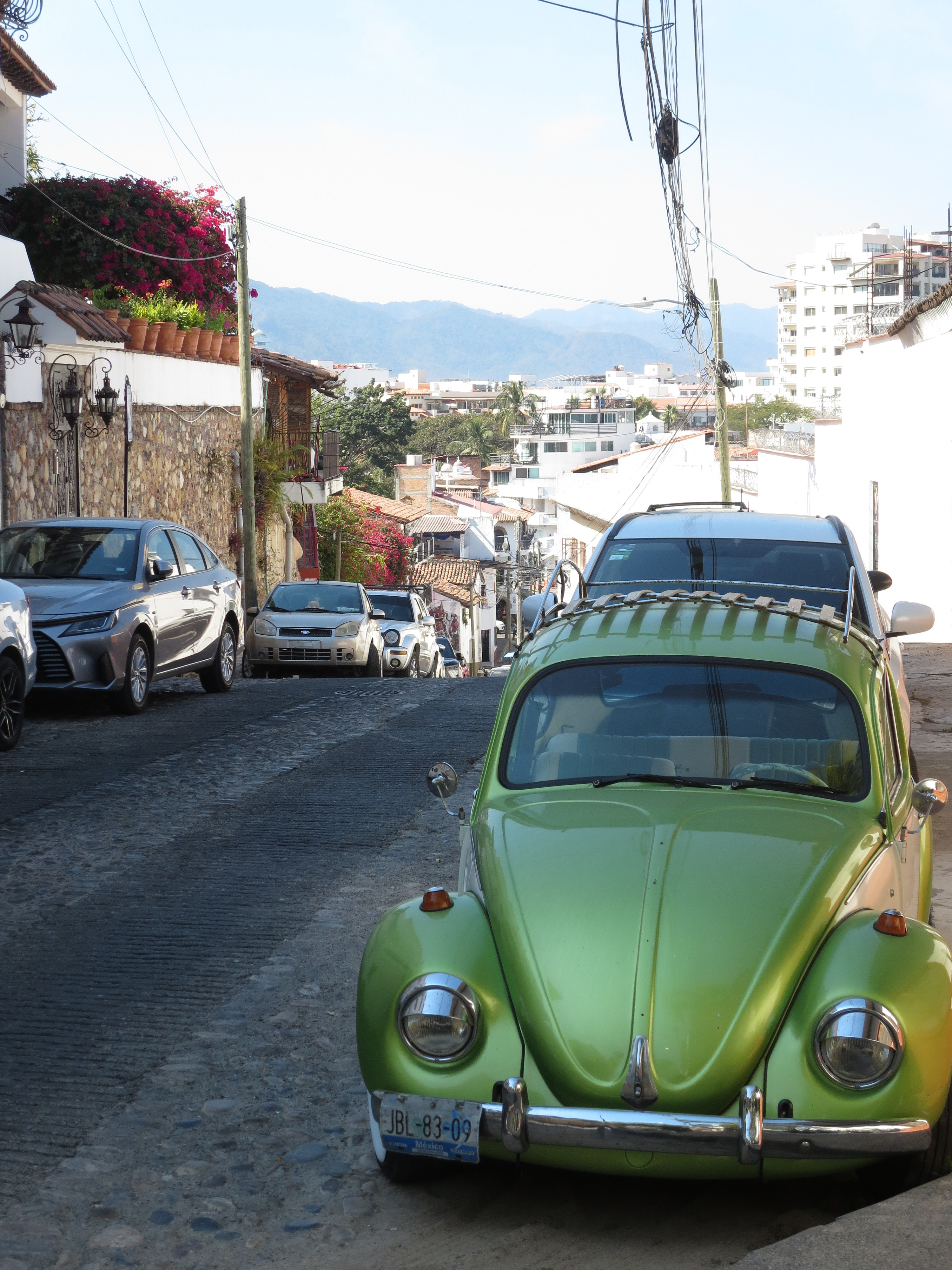Picture facing a downhill stone-brick street. The focus of the image is a shiny dark green VW beetle. While the car is shiny, it is very old and the license plate on the front is rusting.