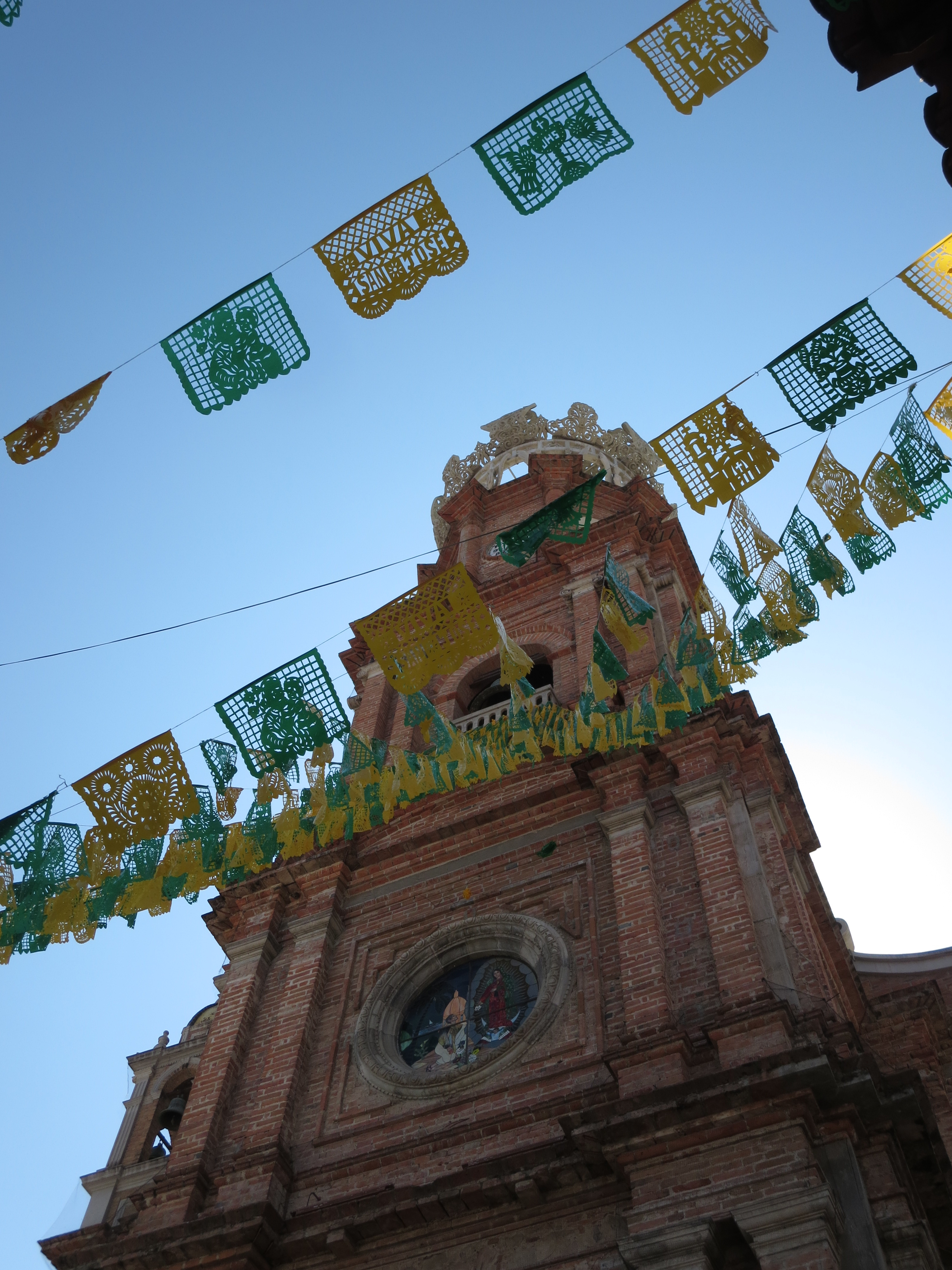 Picture facing towards the sky of a tall church. In between the camera and the church, there are green-and-yellow papeles picados.
