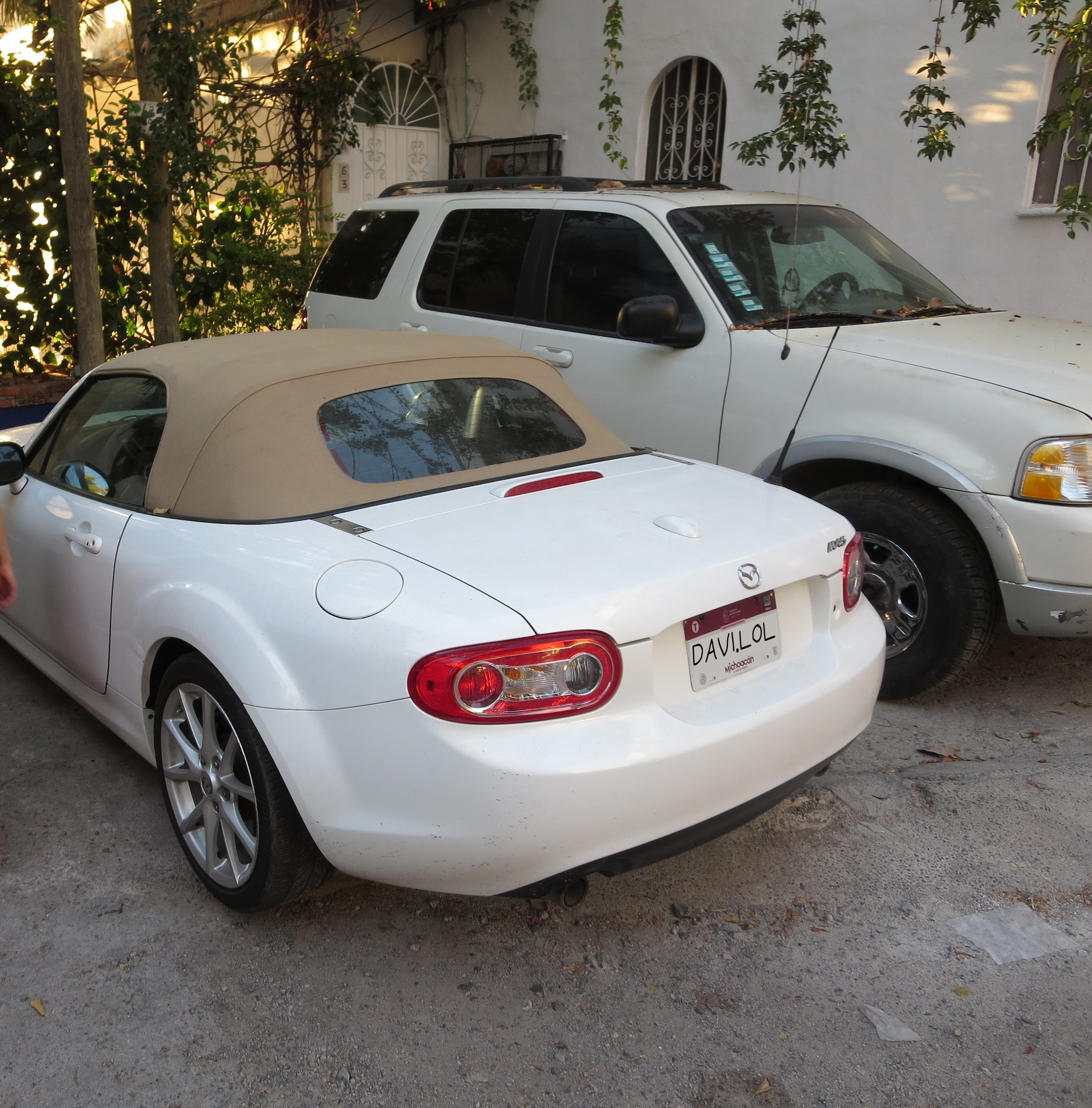 Picture of an NC Mazda MX-5 Miata parked, facing away from the camera next to another car beside a house. License plate is replaced to read DAVI.LOL.